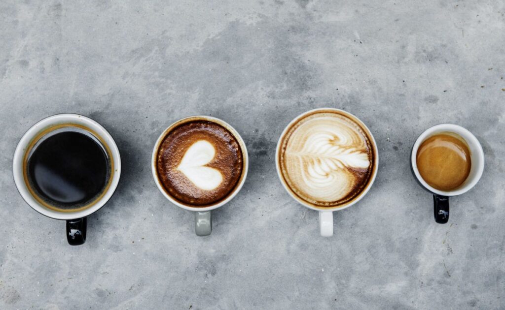 A imagem é uma fotografia tirada na perspectiva de cima de quatro xícaras de café de tamanhos diferentes. Cada xícara contem um tipo de café. A imagem ilustra a tributação do café e da indústria cafeeira. O tema deste artigo.