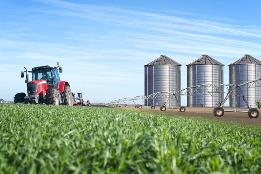 A imagem é a fotografia de um campo de agricultura com um trator a esquerda e três silos a direita. A imagem ilustra a tributação para produtores rurais, o tema desse artigo.