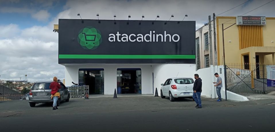 A imagem é a fotografia da entrada do Mercado Atacadinho, localizado no Bairro Alto em Curitiba. A fotografia foi feita durante o dia, é possível ver o banner da fachada do Atacadinho. Na imagem três pessoas aparecem. A foto ilustra o case de sucesso do Mercado Atacadinho, tema deste artigo que comunica a recuperação de créditos tributários para mercados.