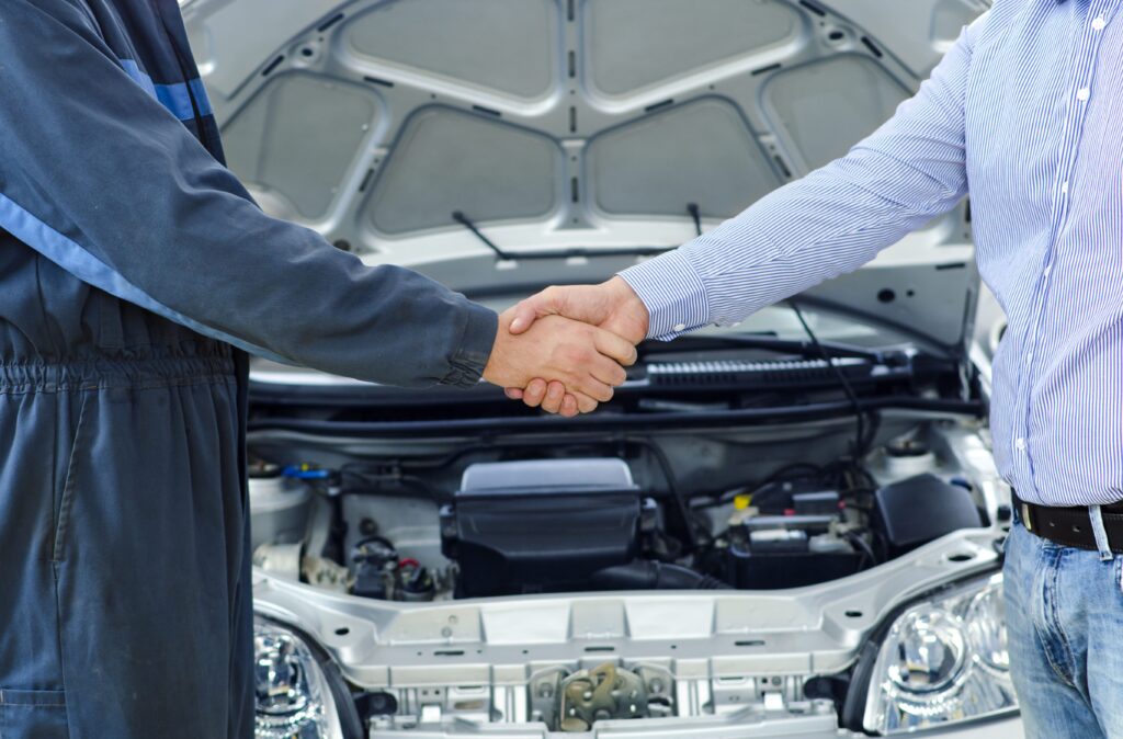 A imagem é a fotografia de um aperto entre duas mãos em frente a um carro com o capô aberto. As mãos são masculinas e possuem a pele branca. A imagem ilustra a recuperação tributária para a indústria automotiva.