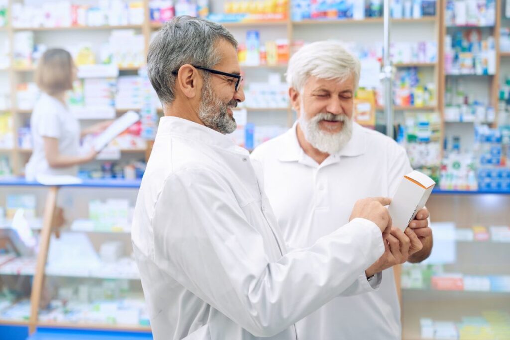 A imagem trata-se da fotografia de dois homens de mais idade olhando  juntos um medicamento em uma farmácia. A imagem representa o fato de que o aumento da população mais 70 é um movimento benéfico para o setor farmacêutico. A foto ilustra o tema de tributação para farmácias.