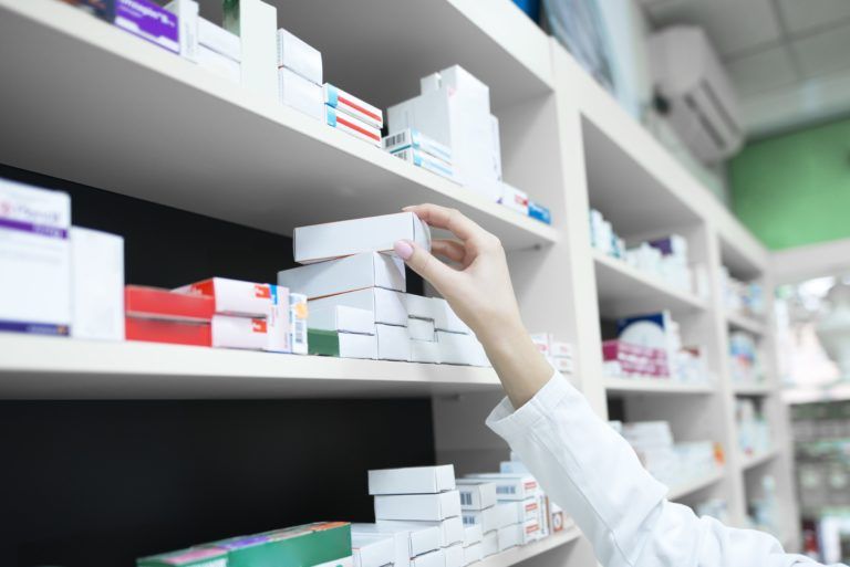A imagem é a fotografia de uma mão de pele branca pegando uma caixa de medicamento em frente a uma estante cheia do produto. É possível ver que o cenário se trata de uma farmácia.