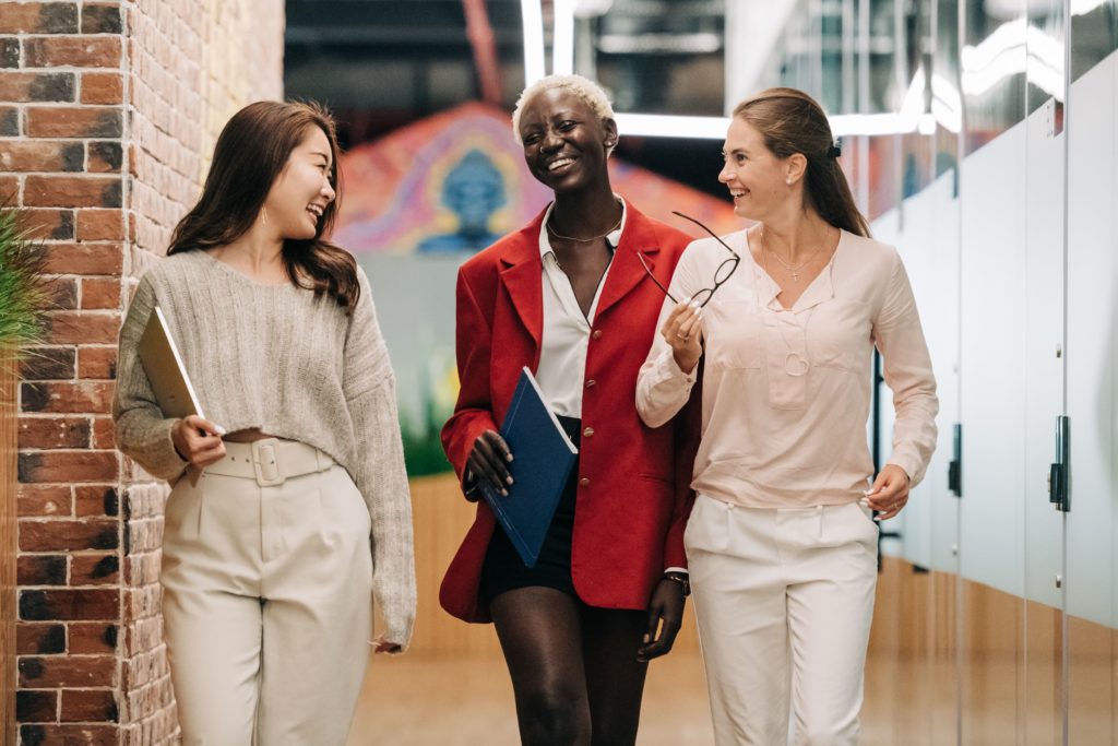 A imagem é a fotografia de três mulheres andando e conversando entre si, todas as três possuem feições alegres em seus rostos. A etnia da primeira mulher é asiática, a segunda é negra e a terceira é branca. As mulheres têm postura e vestem roupas que indicam que elas são empresárias. A foto ilustra a necessidade de conhecer profundamente as obrigações tributárias para atingir o sucesso financeiro em uma empresa.