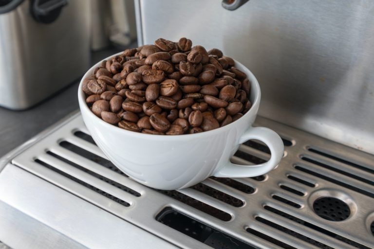A imagem trata-se da fotografia de uma xícara branca cheia de grãos de café. A xícara está apoiada em uma cafeteira. A imagem ilustra a tributação sobre o café, o tema central deste artigo.