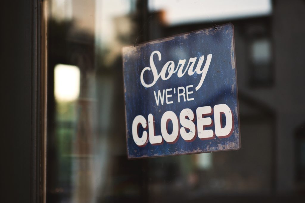 A imagem é a fotografia de uma porta de vidro com uma placa escrito "sorry, we're closed", que em português significa "desculpe, estamos fechados". A imagem ilustra o artigo dicas para evitar falência.