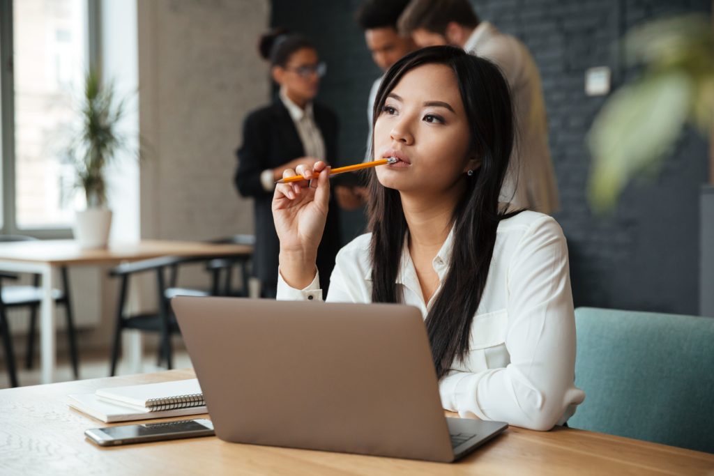 A imagem trata-se da fotografia em cores de uma mulher jovem asiática, ela tem a pele branca e cabelos pretos e lisos. A mulher usa uma blusa de botões branca e está em frente a um notebook. Sua expressão é de dúvida, o que é reforçada pelo fato de ela estar com um lápis na boca, mordendo a ponta com uma expressão pensativa. A foto ilustra o post 3 dicas para evitar a falência da sua empresa.