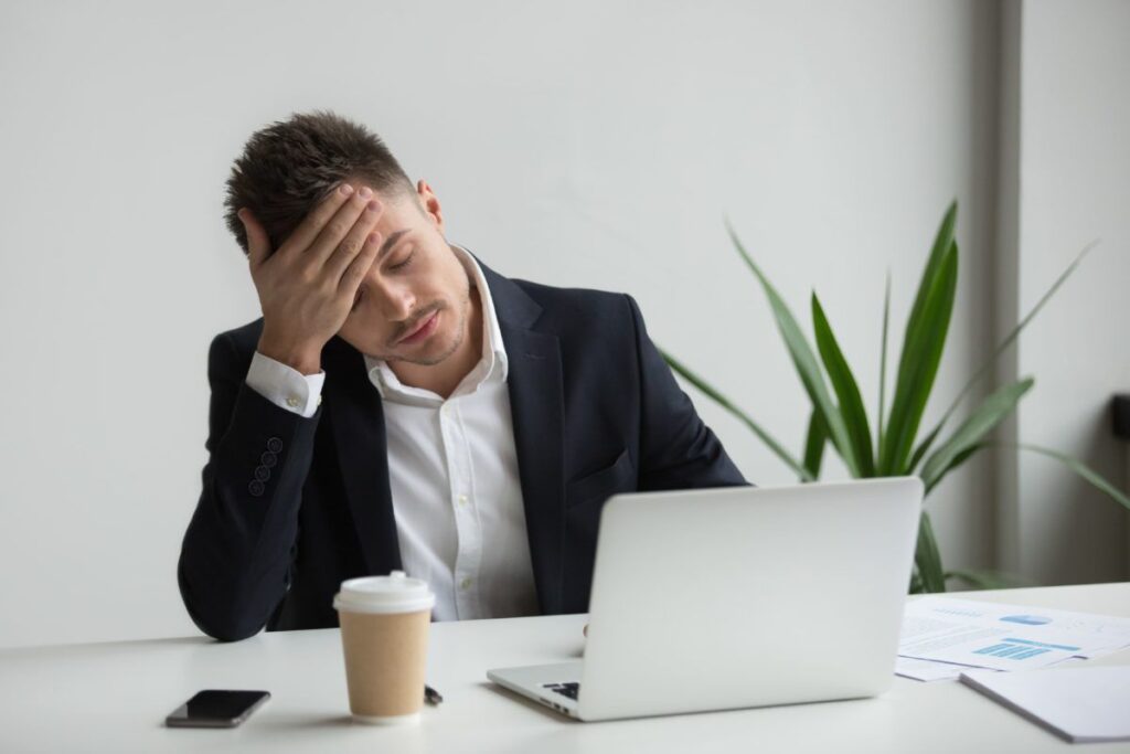 A imagem é a fotografia de um homem branco vestido com um terno em frente a um computador. Ele tem a feição preocupada e está com a mão na cabeça em sinal de apreensão. A imagem ilustra a sonegação de impostos, o tema deste artigo.