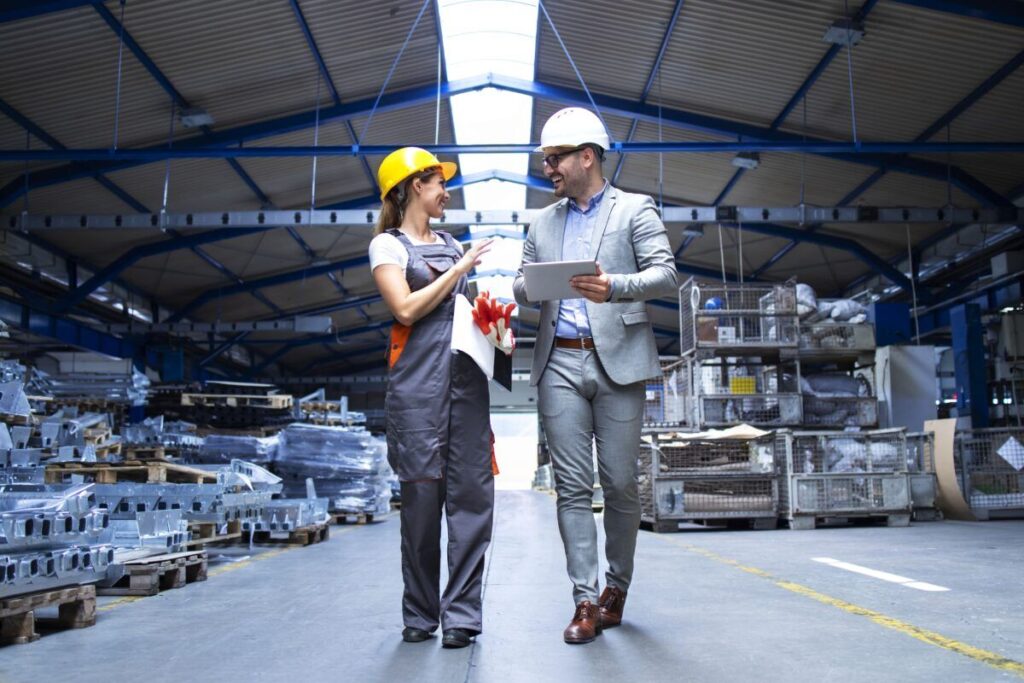 A imagem é a fotografia de dois trabalhadores, um homem e uma mulher em uma fábrica. Ambos tem a pele branca e usam capacetes, o do homem branco e o da mulher amarelo. A mulher veste um macacão em cinza e o homem veste um terno também em cinza. A imagem ilustra a recuperação de tributos sobre insumos.