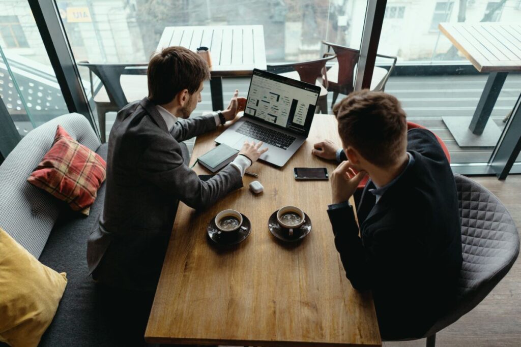 A imagem é a fotografia de dois jovens homens de negócios tendo uma reunião em frente a um notebook. A fotografia é tirada em ângulo plongée. É possível ver que na mesa de reunião tem duas xícaras de café. Ambos os homens têm a pele branca e os cabelos cortados em cortes baixos. A fotografia ilustra os benefícios da recuperação tributária, processo realizado pela Tributo Justo.