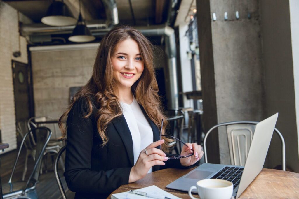 A imagem é a fotografia de um mulher sorridente em uma mesa de trabalho em frente a um notebook. A mulher tem a pele branca, olhos azuis e cabelos claros. Ela veste um blazer azul escuro sobre uma blusa branca. A foto ilustra a necessidade de avaliar se vale a pena fazer a recuperação tributária, o tema deste artigo.