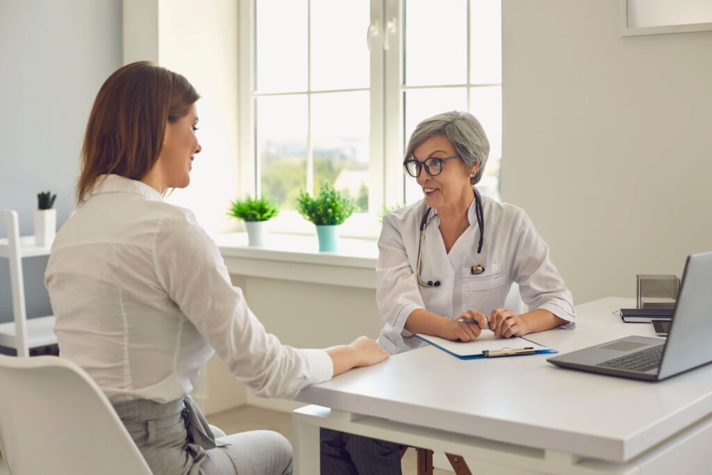 A imagem trata-se da fotografia em cores de duas mulheres - uma médica e uma paciente - conversando entre si em um consultório médico. Ambas as mulheres tem a pele branca, a paciente tem os cabelos castanhos e é um pouco mais jovem do que a mulher médica, que tem cabelos brancos e curtos. A médica tem a expressão feliz em conversar com a paciente, que por estar de costas, tem a face escondida da câmera. 