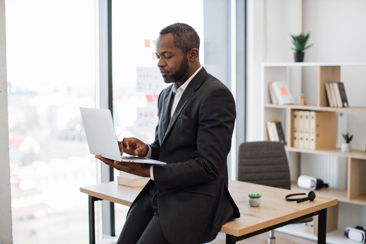 A imagem trata-se da fotografia em cores de um empresário olhando para um notebook, segurado por suas mãos, enquanto está encostado em uma mesa. O homem tem a pele negra, cabelos baixos, e um pouco de barba. Ele veste um terno preto com uma camisa social branca. A foto ilustra este post, cujo tema é gestão tributária.