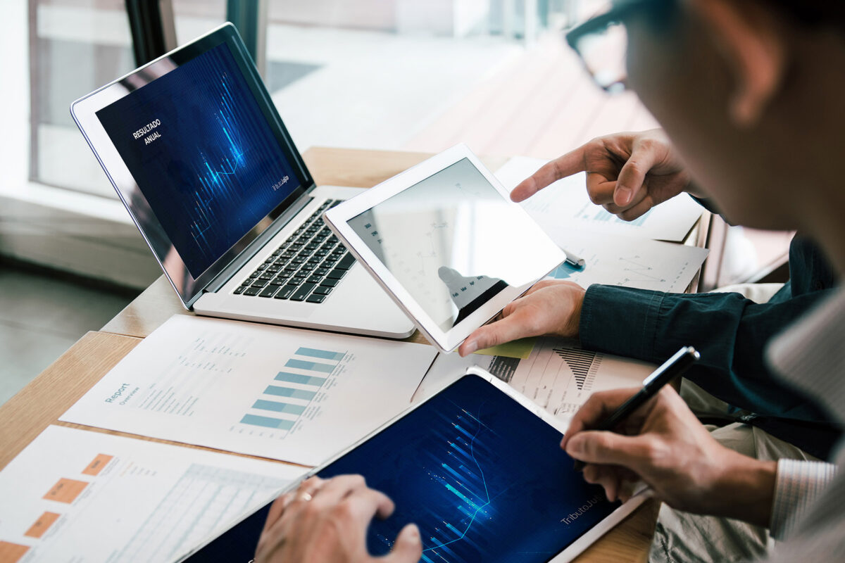 A imagem é uma foto cortada de dois colegas fazendo comparações em um notebook e tablets de gráficos de saúde financeiros.